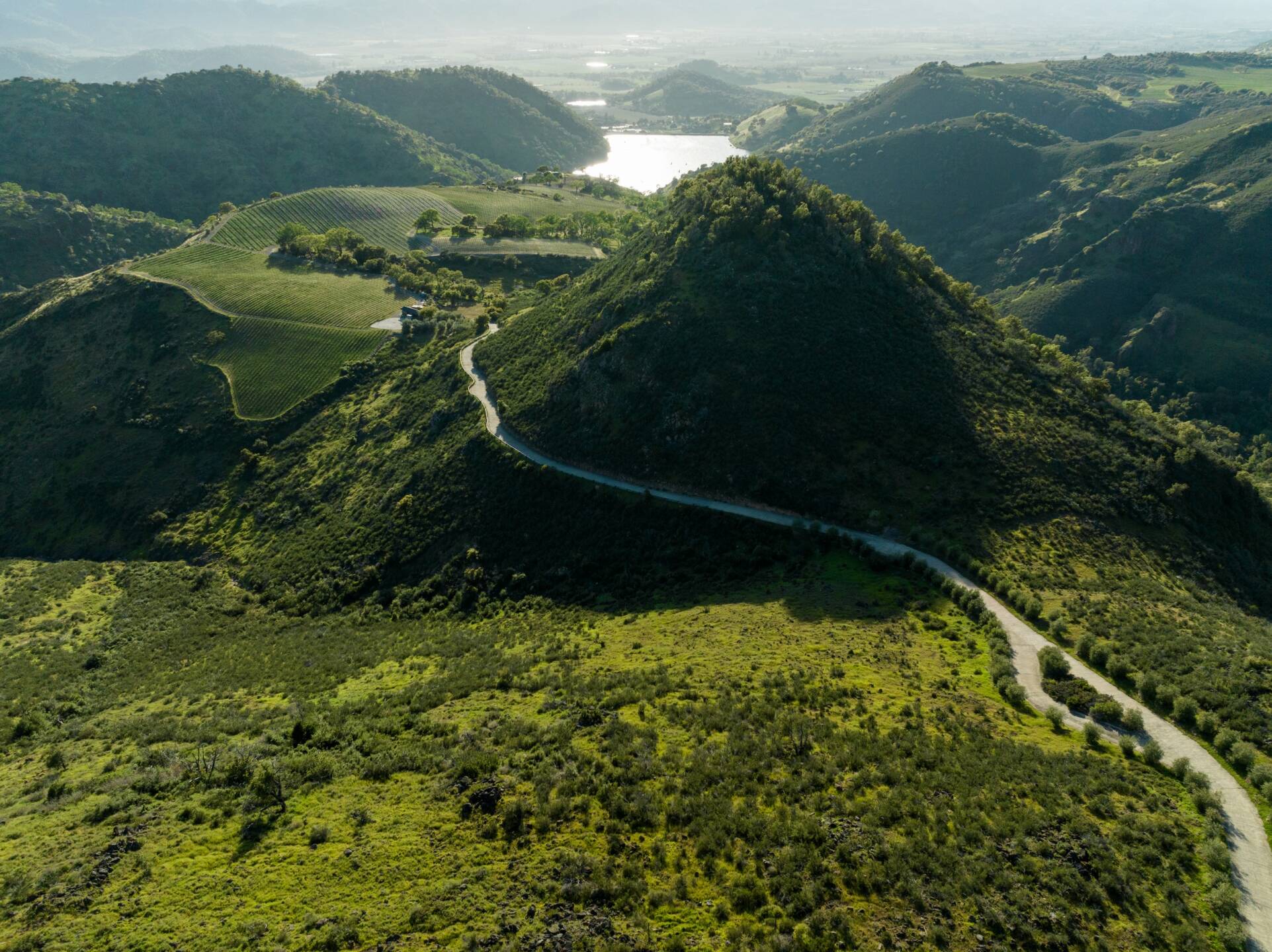 A winding road snakes through green hills with a large body of water visible in the distance under a partly cloudy sky.