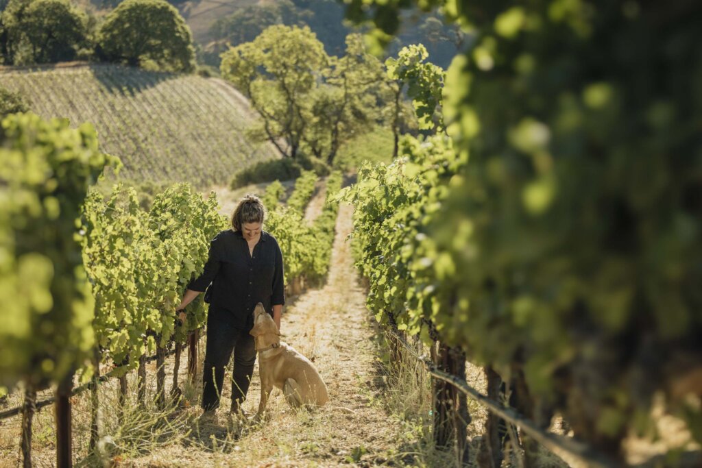 Winemaker Katie Vogt walking with a dog through Waters Ranch vineyard with green grapevines and rolling hills in the background.