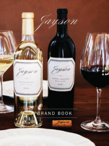 Two bottles of Jayson wine, one Sauvignon Blanc and one Cabernet Sauvignon with wine glasses and a cork on a table set for dining. Text reads "BRAND BOOK.