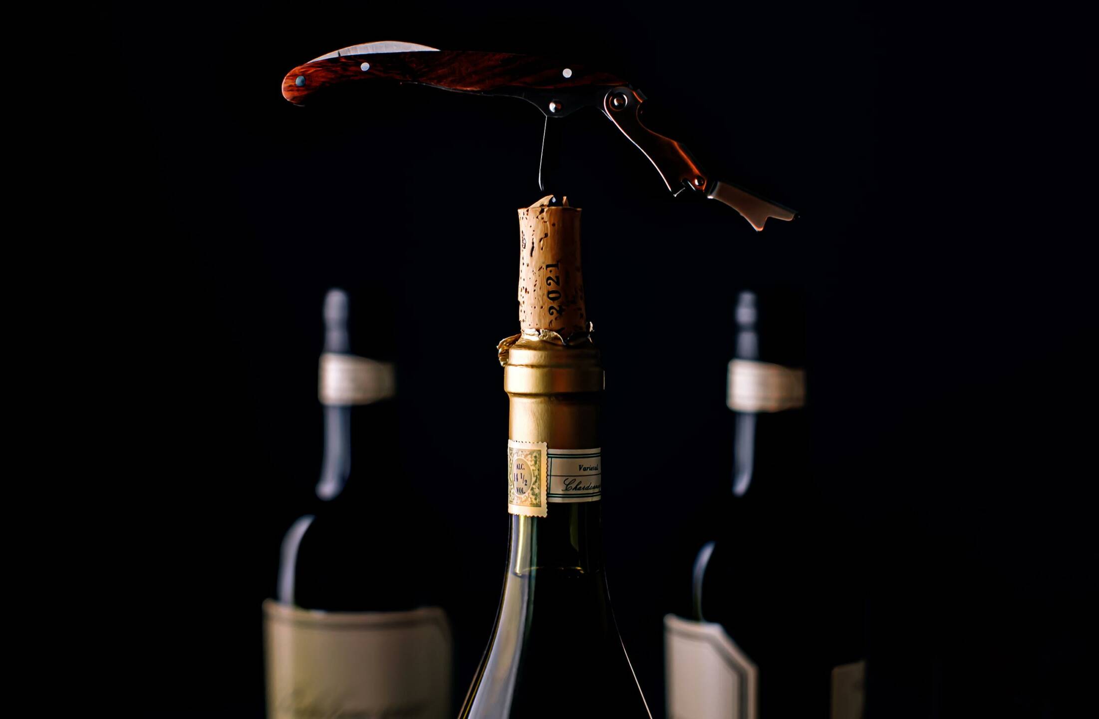 A corkscrew is inserted into the cork of a wine bottle with two more blurred wine bottles in the background against a dark backdrop.