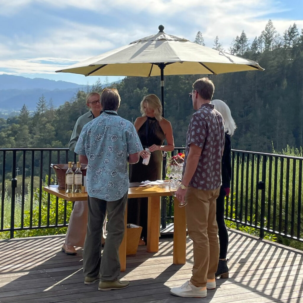 Five people standing around a table under an umbrella are tasting wine on an outdoor deck with a scenic mountain view in the background.