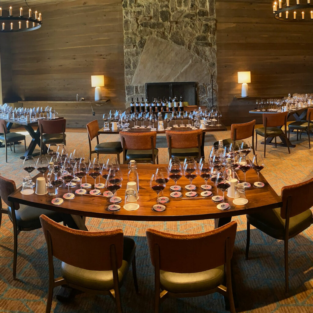 A room set up for a wine tasting event. Multiple tables hold numerous wine glasses and coasters, with a stone fireplace and warm lighting in the background.