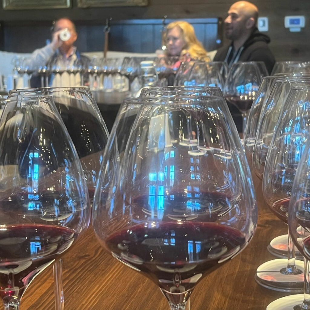Several glasses of red wine are arranged on a table with three people in the background, possibly at a wine tasting event. The background is slightly blurred, making the wine glasses the focal point.
