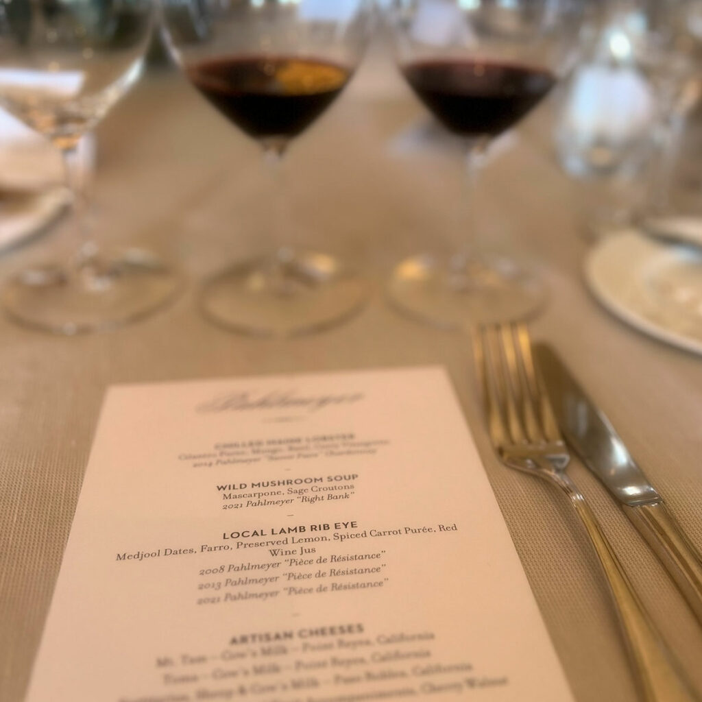 Table setting with a white tablecloth, two glasses of red wine, and a menu listing dishes such as wild mushroom soup, local lamb rib eye, and artisan cheeses. Knife and fork placed beside the menu.