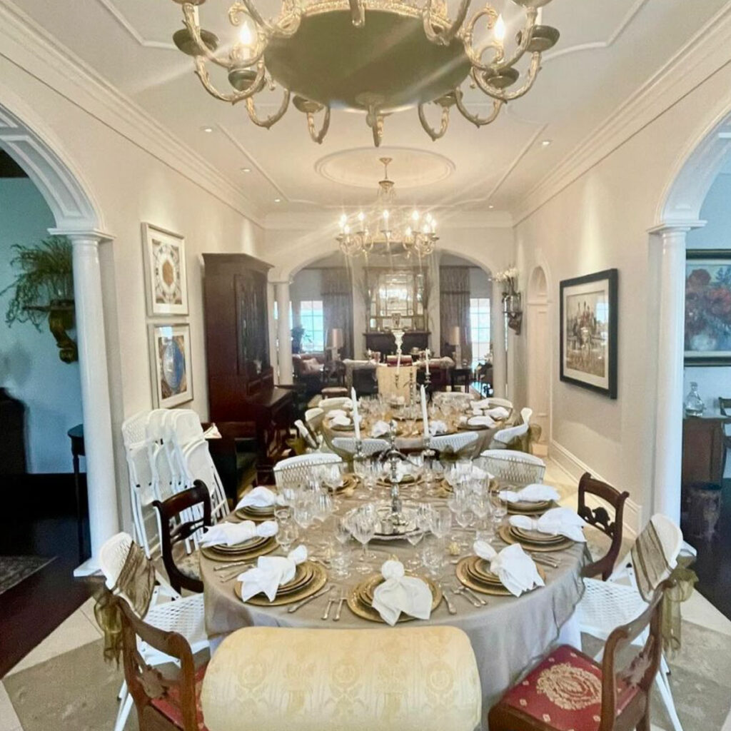 A dining room with a long table set for a meal, featuring plates, wine glasses, and cloth napkins. The room is adorned with chandeliers, framed artwork, and elegant decor.