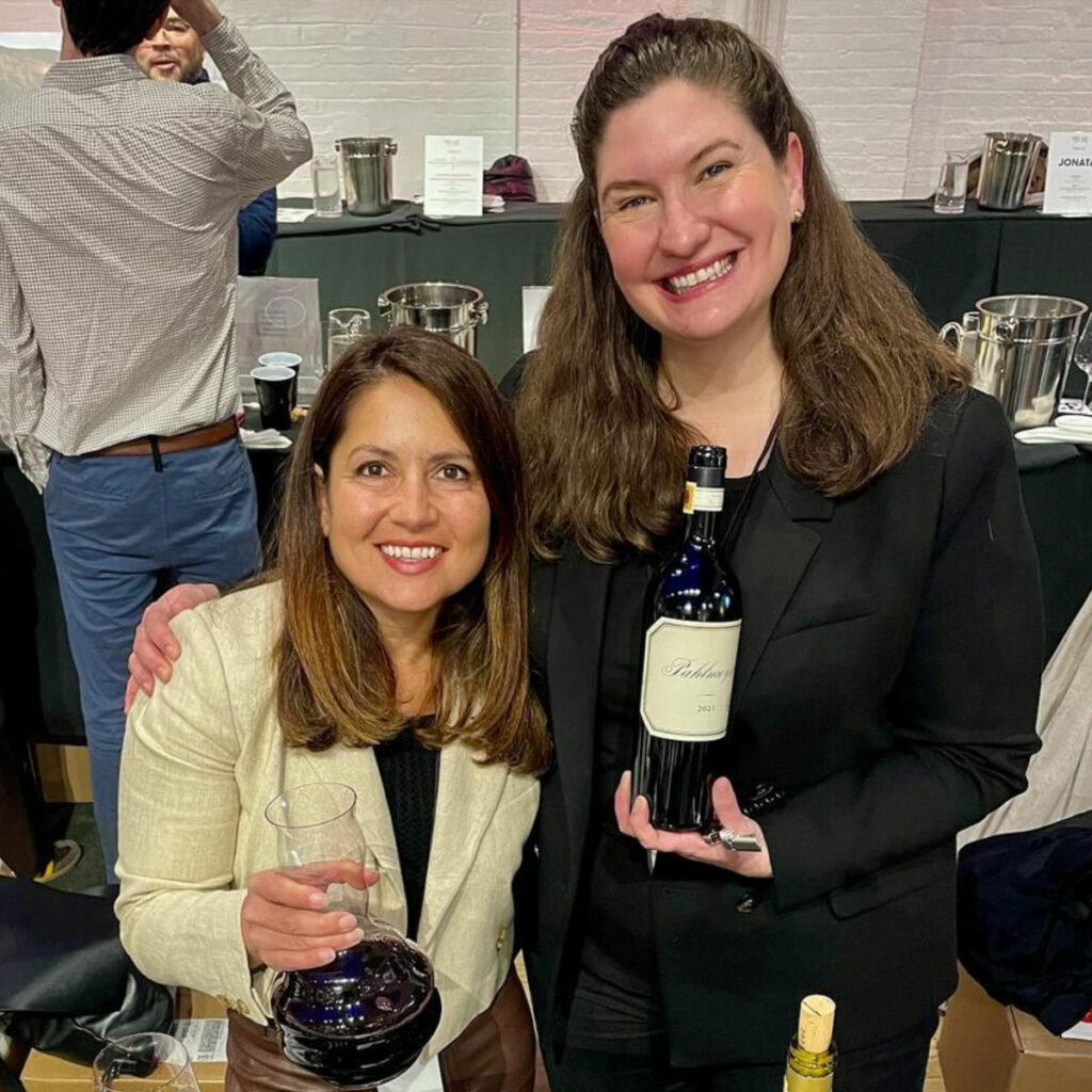 Two women smiling and holding wine at a tasting event, with one holding a bottle of wine and the other holding a glass filled with red wine.