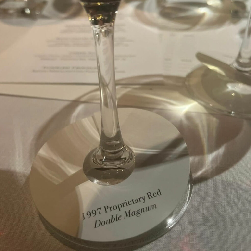 Close-up of a wine glass on a coaster imprinted with "1997 Proprietary Red Double Magnum" on a white tablecloth with a blurred background.