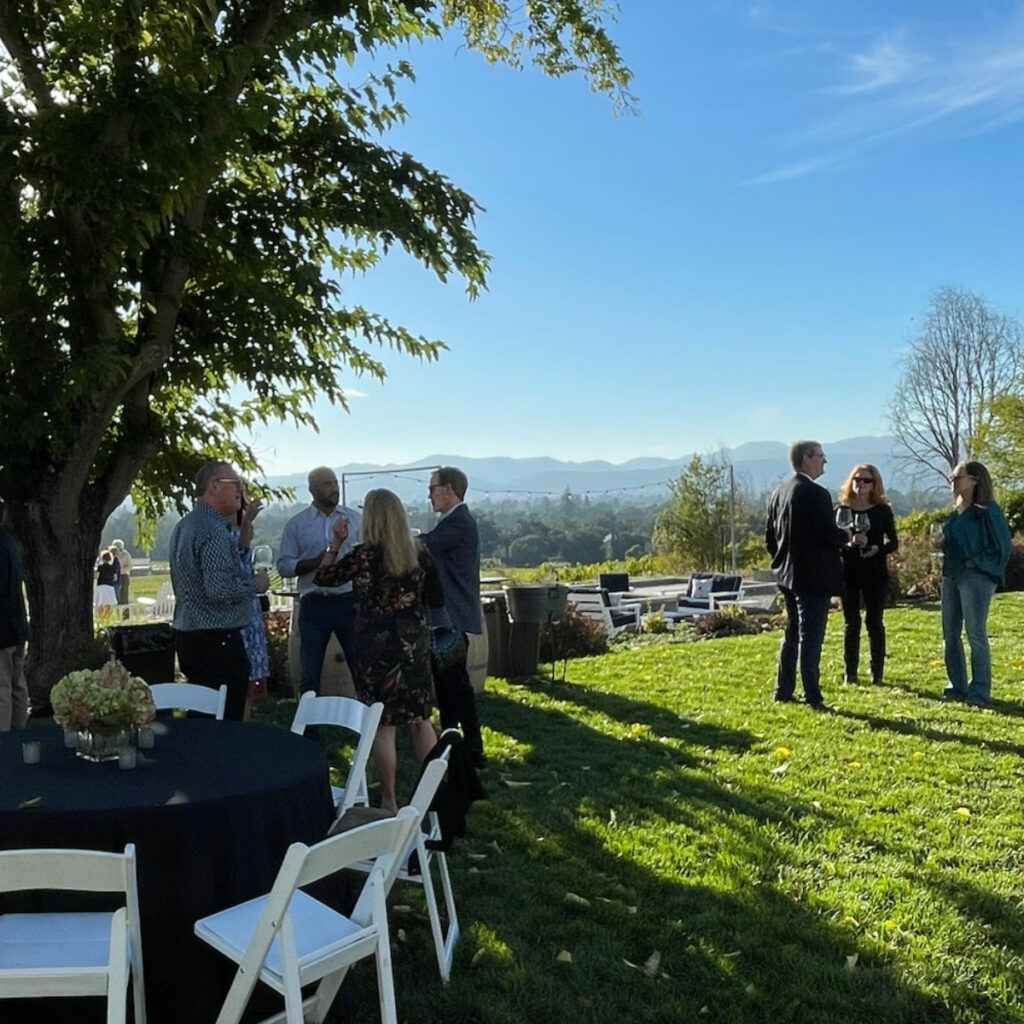 People gather and converse outdoors on a grassy area under clear skies, with a scenic backdrop of mountains and trees. White chairs and a table with a floral centerpiece are visible in the foreground.
