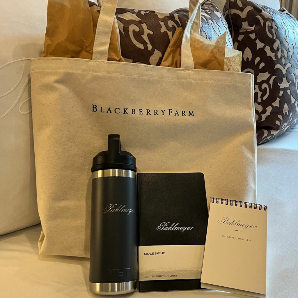 A tote bag labeled "Blackberry Farm" containing a stainless steel water bottle, a Moleskine notebook, a smaller notepad, and some tissue paper sits on a cushioned seat.