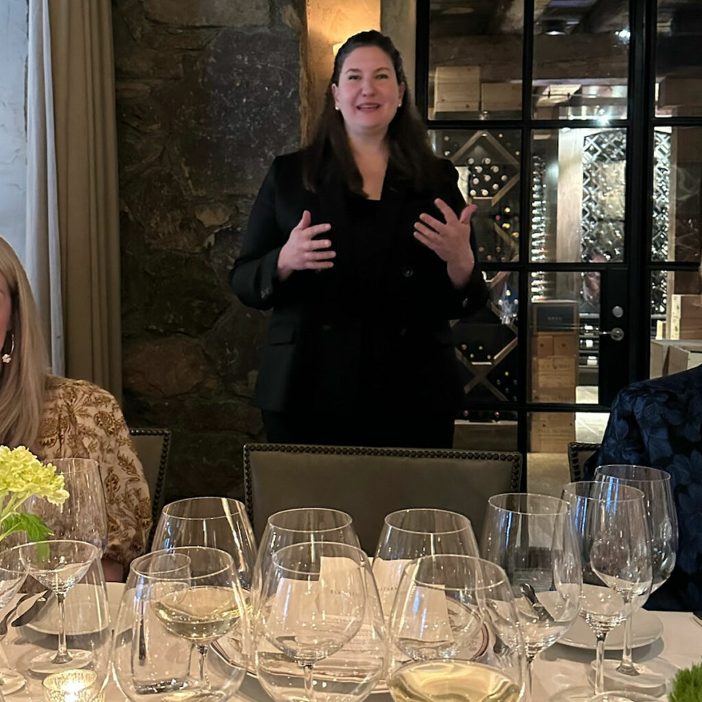 A woman stands and gestures with her hands, speaking in a dining room with wine glasses on the table in the foreground.