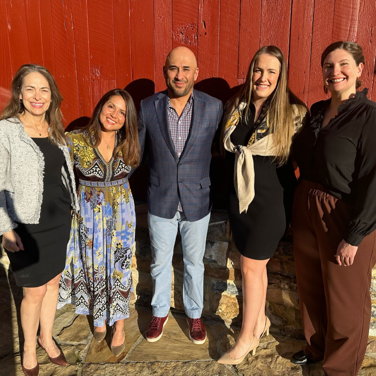 Five people are standing in a row outdoors in front of a red wall. They are all smiling and dressed in formal attire, and the sunlight casts shadows on the ground.