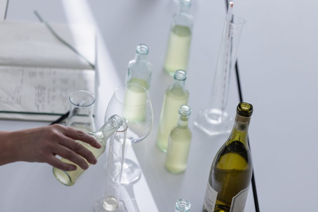 A hand pours liquid from a bottle into a glass on a table. Various bottles, beakers, and an open book are also on the table.