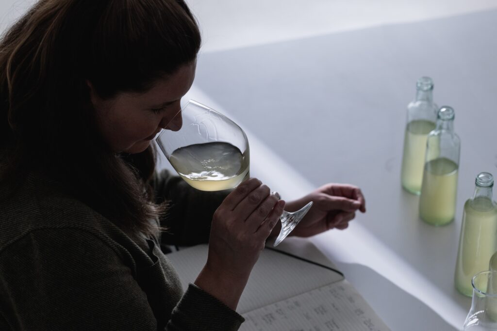 A person smelling white wine in a glass while seated at a table with a notebook and three bottles of liquid.