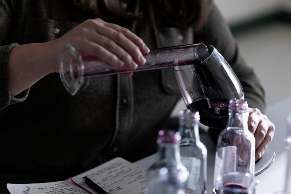 A person pours red wine from a test tube into a glass, with several bottles and a notebook visible on the table.