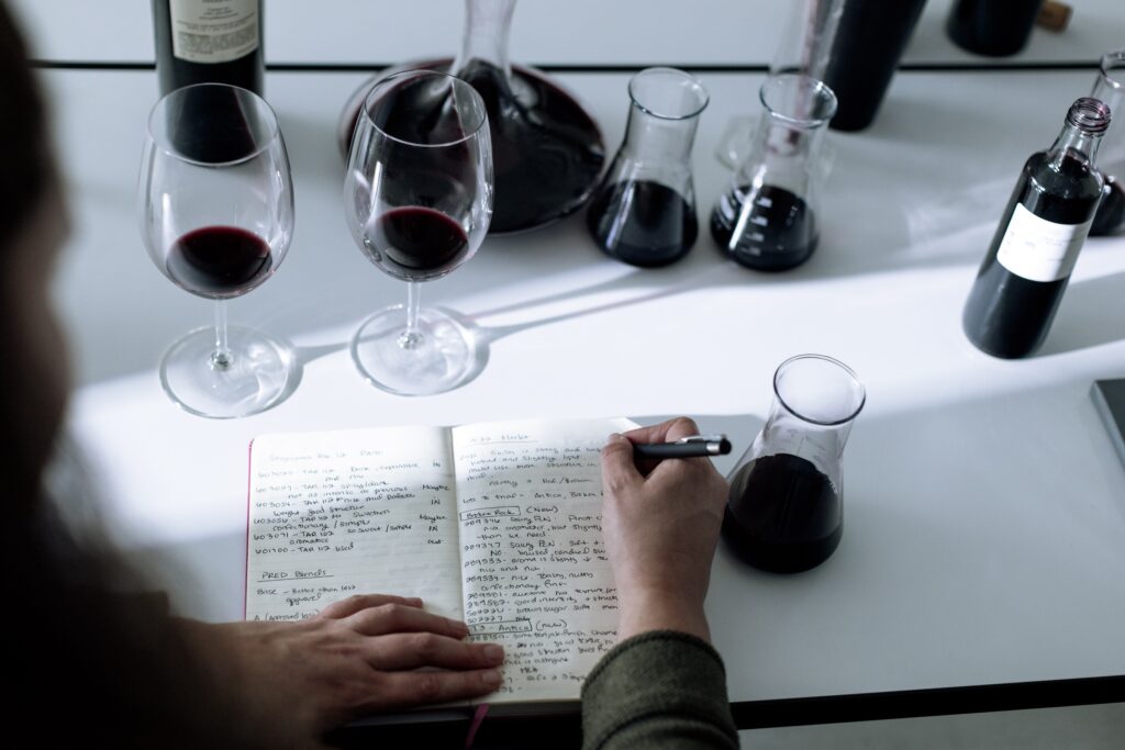 Person writing notes in a journal surrounded by carafes and glasses of red wine on a table.