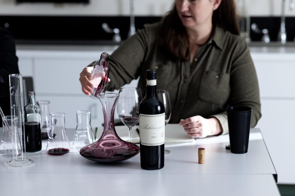 A person pours red wine from a bottle into a decanter on a table with glasses and beakers.