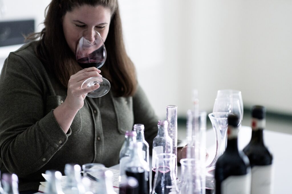 Woman smelling a glass of red wine at a table with various wine bottles and glasses.