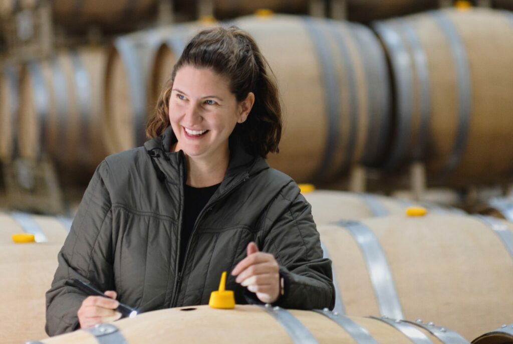 A person smiles while working in a winery, surrounded by stacks of wine barrels.