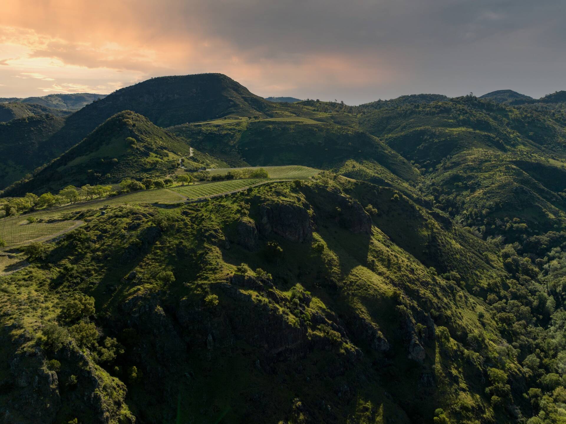 Aerial view of a sprawling green mountain range under a partly cloudy sky during sunset.