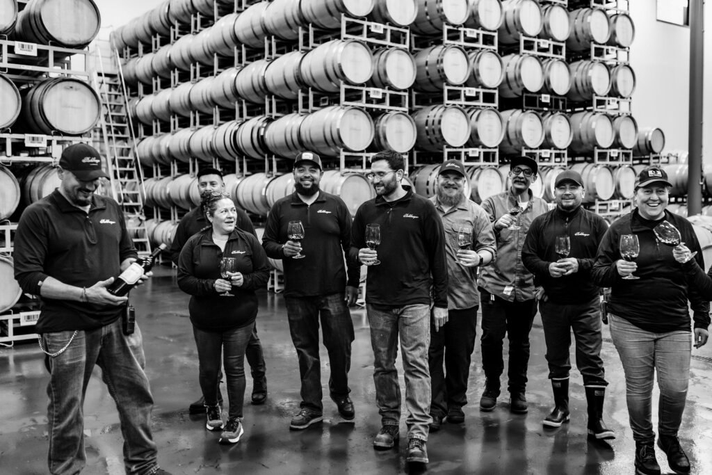 A group of eight people stand in a winery with wine glasses, in front of stacks of barrels. Some are looking at the camera, and one person is holding a wine bottle.