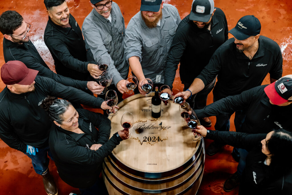 A group of people stand around a wooden barrel, toasting with glasses of red wine. The barrel is inscribed with "Vintage Robert Mondavi 2024.