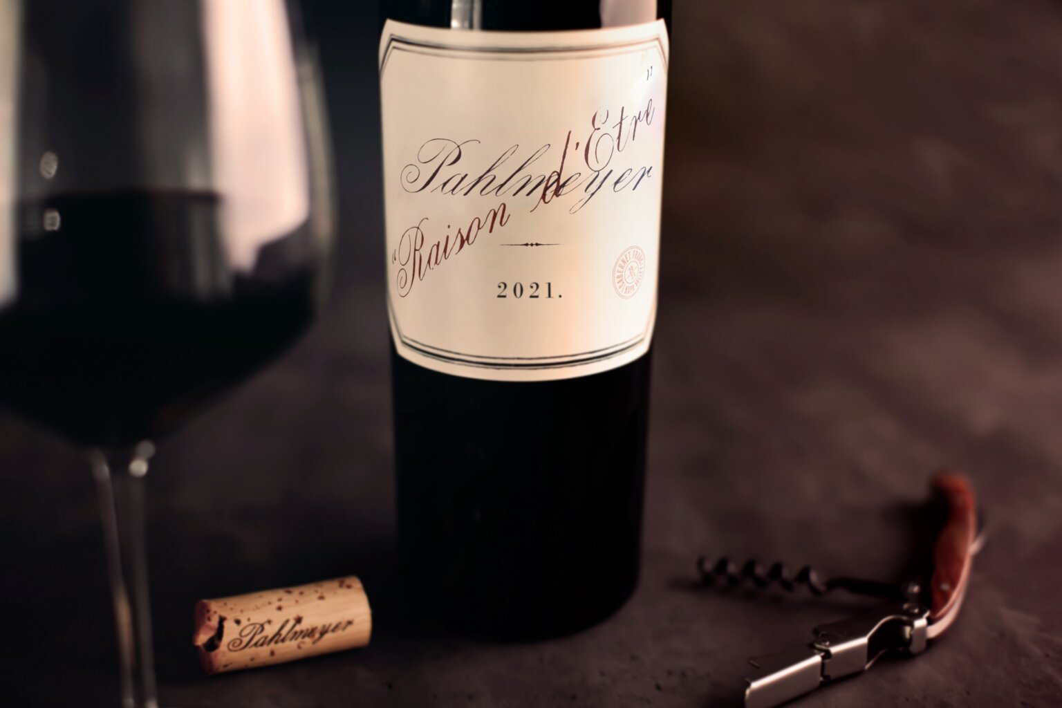 A bottle of 2021 vintage wine labeled "Raison d'être" next to a wine glass, a cork, and a corkscrew on a dark surface.